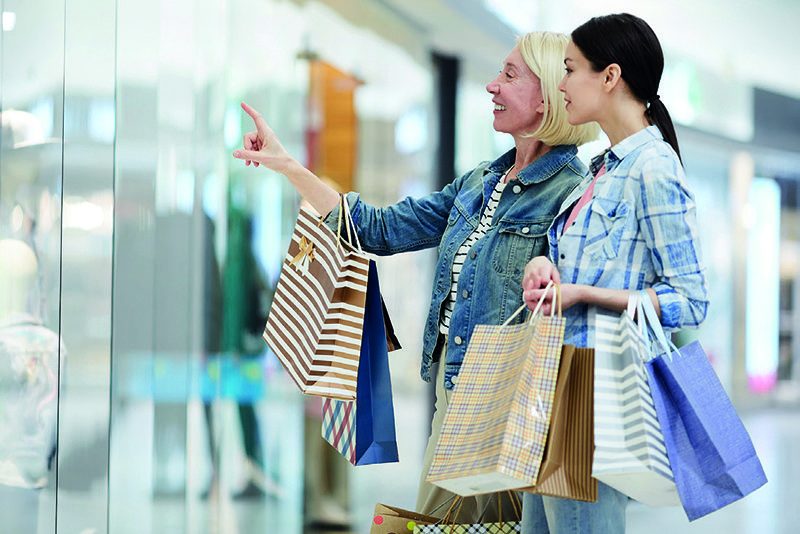 Cheerful mother and daughter discussing clothing of mannequins Cheerful excited mature mother and adult daughter in casual clothing standing in corridor of mall and discussing clothing of mannequins