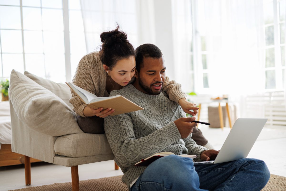Young asian woman with book supporting to african american boyfriend with online courses while using laptop together at home